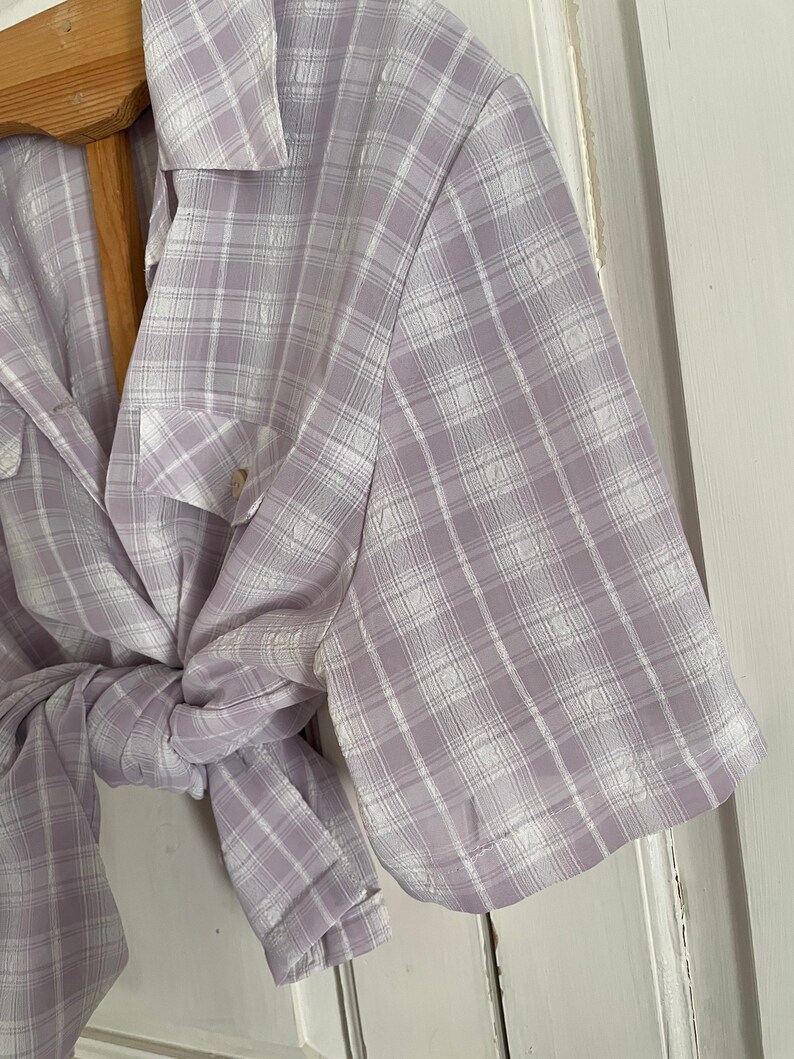 May include: A light purple and white plaid shirt with short sleeves and a button-up collar. The shirt is tied at the waist.