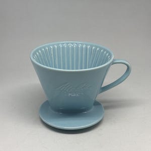 Könnte beinhalten: Ein hellblauer Keramik-Kaffeefilter mit Griff und kleinem Sockel. Der Filter hat vertikale Rillen auf der Innenseite und der Markenname "Melitta" ist auf der Vorderseite eingeprägt. Dies ist ein klassischer Pour-Over-Kaffeebereiter.