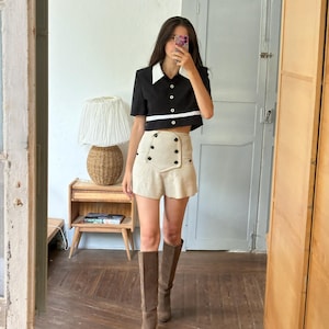 May include: A black short-sleeved collared shirt with a white collar and a horizontal white stripe across the midsection, paired with high-waisted beige shorts featuring a double-breasted button detail and brown knee-high suede boots.