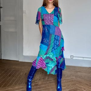 Pode incluir: Um vestido midi com um design de patchwork de painéis de tecido azul, turquesa e roxo com vários padrões florais e geométricos. O vestido tem um decote em V, mangas curtas esvoaçantes e uma bainha com folhos, combinado com botas azuis brilhantes.