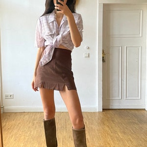 May include: A woman wearing a light purple plaid shirt tied at the waist, a brown mini skirt with side slits, and brown knee-high boots.