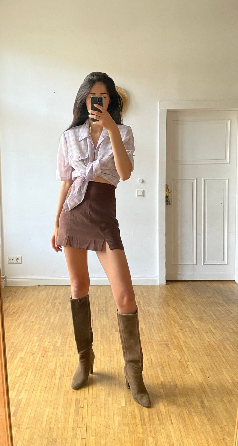 May include: A woman wearing a light purple plaid shirt tied at the waist, a brown mini skirt with side slits, and brown knee-high boots.
