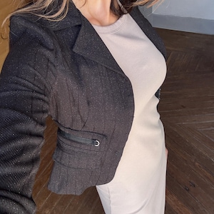 Peut inclure: Un blazer noir avec un motif texturé et un petit détail de poche. Le blazer est associé à une robe beige clair. La photo est prise d'un angle légèrement surélevé, mettant en valeur les textures et la coupe des vêtements.