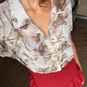 Puede incluir: Una camisa de manga corta con botones y estampado floral en tonos marrón y beige. Combinada con una minifalda roja con volantes. La imagen se toma desde arriba, destacando las texturas y colores de la ropa.