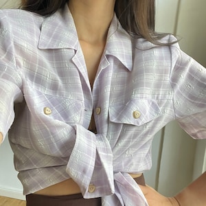 May include: A light purple plaid button-down shirt with short sleeves. The shirt is tied at the waist and has two flap pockets with buttons.