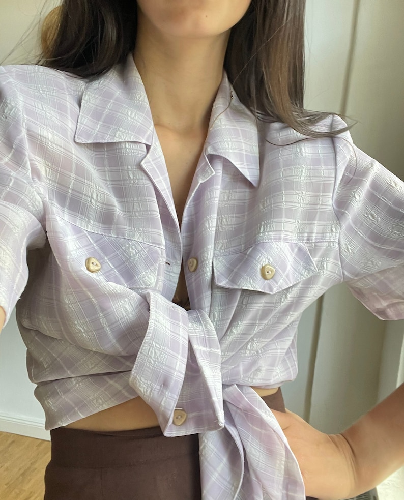 May include: A light purple plaid button-down shirt with short sleeves. The shirt is tied at the waist and has two flap pockets with buttons.