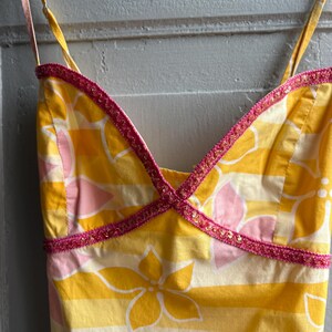 May include: A yellow sundress with a floral pattern and pink accents. The dress features a V-neckline trimmed with pink sequined trim and thin spaghetti straps. The fabric has horizontal stripes and floral designs.