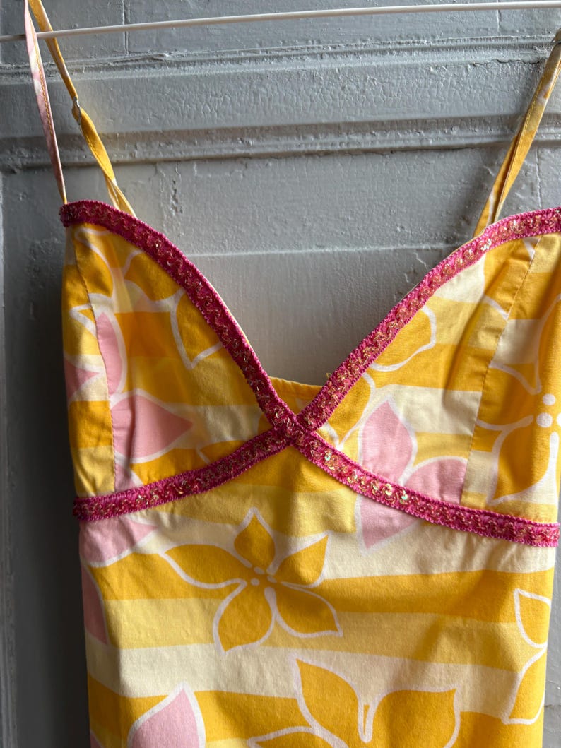 May include: A yellow sundress with a floral pattern and pink accents. The dress features a V-neckline trimmed with pink sequined trim and thin spaghetti straps. The fabric has horizontal stripes and floral designs.