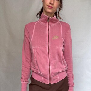 K&ouml;nnte beinhalten: Rosafarbene Jacke mit Rei&szlig;verschluss, hohem Kragen und zwei Vordertaschen. Die Jacke hat das goldene Nike-Logo auf der Brust. Die &Auml;rmel und Schultern haben eine gerippte Textur. Die Jacke wird mit einem braunen Rock getragen.