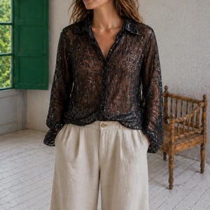 Può includere: Una camicia nera trasparente con bottoni, maniche lunghe e colletto, ornata da paillettes scintillanti. Abbinata a pantaloni beige chiaro a gamba larga. L'ambientazione è in una stanza con una sedia di legno e una finestra verde.