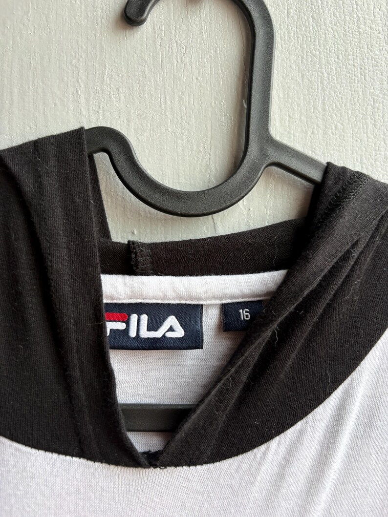 May include: A white and black hooded sweatshirt with the brand name "FILA" in white letters on a navy blue label with a red stripe. The size "16" is printed on a separate label.