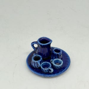 Peut inclure: Un service à thé miniature en céramique bleue, comprenant une petite cruche, quatre tasses et un plateau rond. L'ensemble est d'un bleu profond et vibrant, avec une finition brillante. Les tasses ont de petites anses et un motif côtelé.