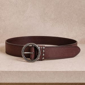 Pode incluir: Um cinto castanho escuro com uma fivela circular de metal prateado texturizado. A tira do cinto é feita de um material liso com costuras visíveis nas bordas e inclui uma fila de quatro pequenos rebites de metal prateado perto da fivela.
