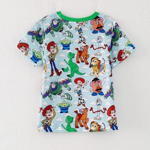 K&ouml;nnte beinhalten: Hellblaues T-Shirt mit einem farbenfrohen Muster von Figuren aus dem Animationsfilm Toy Story. Das Shirt hat einen gr&uuml;nen Halsausschnitt und kurze &Auml;rmel. Das Design umfasst Bilder von Woody, Buzz Lightyear und anderen Charakteren.