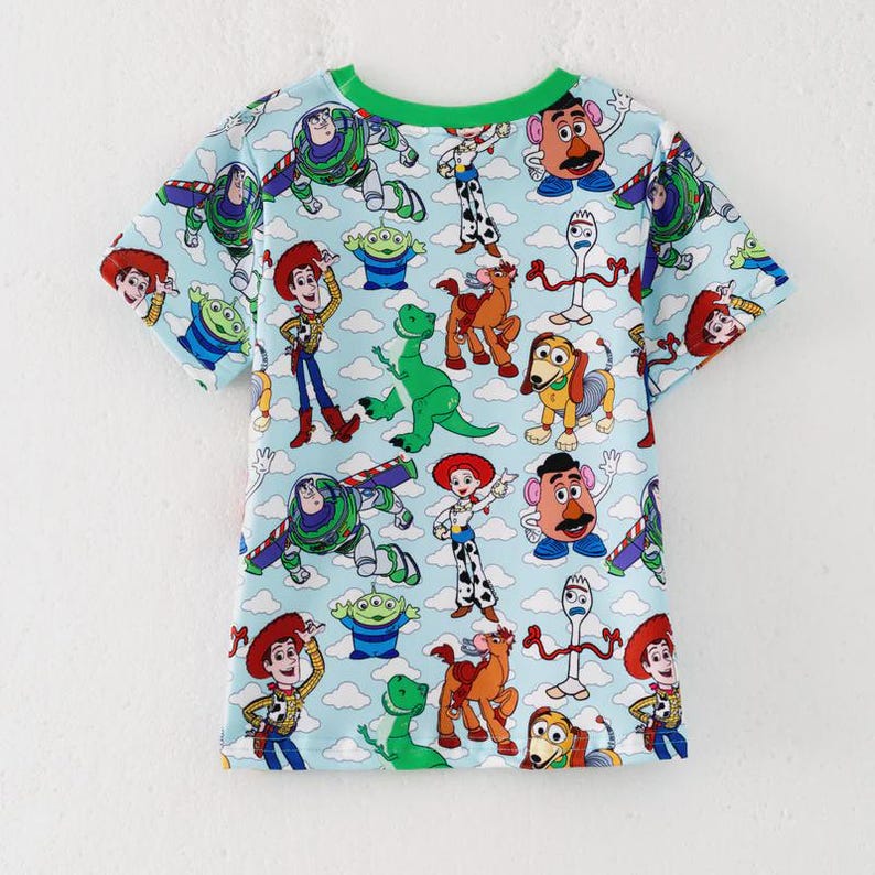 K&ouml;nnte beinhalten: Hellblaues T-Shirt mit einem farbenfrohen Muster von Figuren aus dem Animationsfilm Toy Story. Das Shirt hat einen gr&uuml;nen Halsausschnitt und kurze &Auml;rmel. Das Design umfasst Bilder von Woody, Buzz Lightyear und anderen Charakteren.