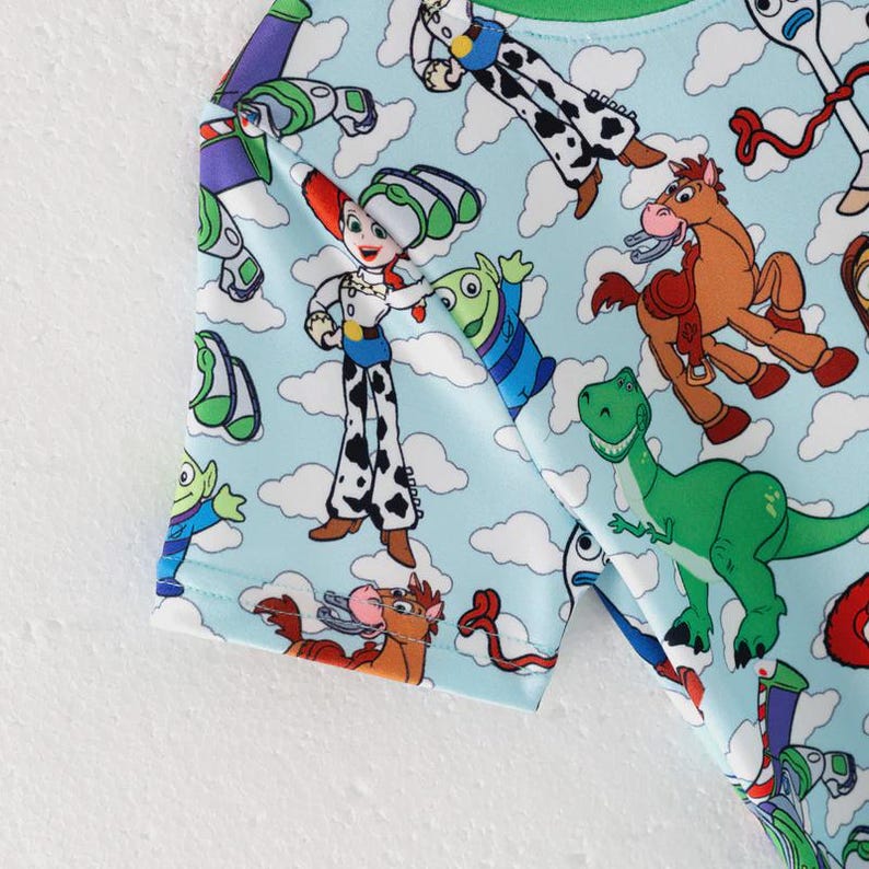 K&ouml;nnte beinhalten: Hellblaues T-Shirt mit einem farbenfrohen Aufdruck von Toy Story-Charakteren. Das Design zeigt Woody, Buzz Lightyear, Jessie, Rex und andere Charaktere vor einem Hintergrund aus wei&szlig;en Wolken. Das T-Shirt ist aus weichem Stoff.