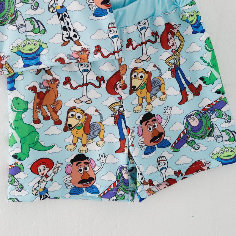 K&ouml;nnte beinhalten: Hellblaue Shorts mit einem sich wiederholenden Muster von Toy Story-Charakteren wie Woody, Buzz Lightyear und Mr. Potato Head. Das Design umfasst Wolken und verschiedene Charaktere aus dem Film. Die Shorts haben einen hellblauen Bund.