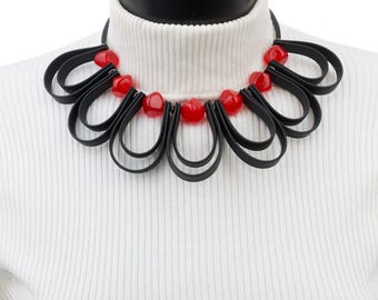 May include: A black and red statement necklace with multiple black loops and red beads. The necklace is displayed on a white turtleneck sweater.