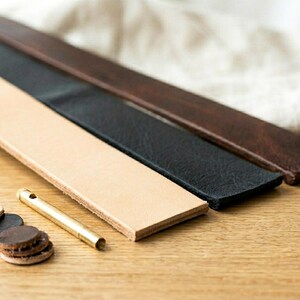 May include: Three leather strips in tan, black, and brown, with a brass tube and several small leather circles. The leather strips are arranged diagonally on a wooden surface, with a white cloth in the background.