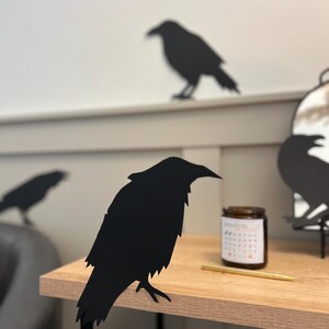Puede incluir: Decoración de silueta de cuervo negro sobre una mesa de madera con un soporte de metal negro. El cuervo está posado en la mesa con las alas extendidas.