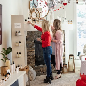 May include: Two people are standing in front of a fireplace, looking at a shelf of small bottles labelled "Herbal". The bottles are arranged in rows on a white shelf. The fireplace is made of stone and has a wooden mantel. There are candles and other decorations on the mantel.