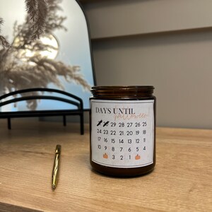 Könnte beinhalten: Ein braunes Glasgefäß mit einem weißen Etikett, auf dem "DAYS UNTIL halloween" steht und ein Countdown der Tage bis Halloween abgebildet ist. Das Gefäß steht auf einer Holzoberfläche mit einem goldenen Stift daneben.