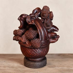 May include: A dark brown carved wooden sculpture of a basket filled with textured fruit and flowers. The basket features a woven lattice pattern on the exterior and a large, smooth handle arching over the top, resting on a circular base.