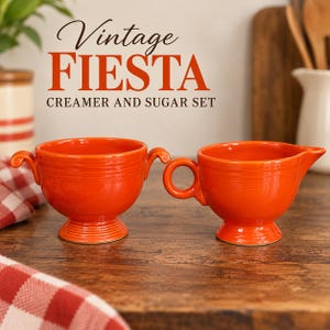 May include: A bright orange ceramic creamer and sugar bowl set with a glossy finish. The sugar bowl features two side handles, while the creamer has a single handle and a pouring spout. Both pieces have a pedestal base and embossed ring details.