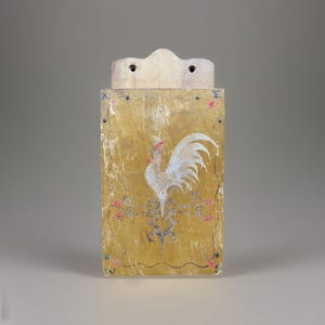Op de afbeelding: Een rechthoekige houten wandzak met een verweerde mosterdgele afwerking. De voorkant is voorzien van een geschilderde witte haanillustratie, decoratief krulwerk en de letters W, E en N in de hoeken. Het heeft twee gaten aan de bovenkant voor montage.
