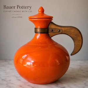 May include: A bright orange Bauer Pottery coffee carafe with a matching lid, featuring a curved wooden handle attached with a brass-colored metal band. The text Bauer Pottery, COFFEE CARAFE WITH LID, and circa 1930 is printed above the carafe.