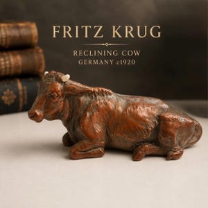 May include: A brown, reclining cow figurine with small white horns. The text above the object reads: FRITZ KRUG, RECLINING COW, GERMANY c1920. The sculpture features a textured surface and is positioned in a resting pose with legs tucked under.