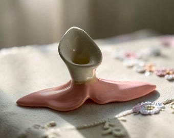 Könnte beinhalten: Ein Kerzenhalter aus Keramik mit einem glatten, cremefarbenen Oberteil in Form einer Calla-Lilienblüte und einem breiten, flachen, blassrosa Sockel, der sich in einer fließenden, organischen Form ausbreitet.