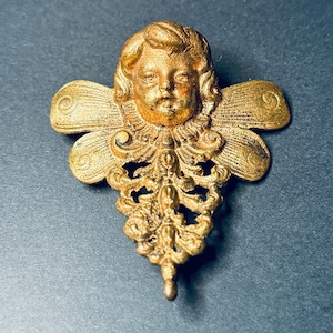 May include: A gold-toned metal brooch featuring a cherub face at the center with stylized wings extending to the sides. Below the face, an intricate, ornate filigree design tapers downward to a point, creating a symmetrical decorative piece.