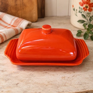 May include: A bright orange ceramic butter dish with a rectangular base and a matching lid. The lid features a small, centered knob handle, while the base has ribbed edges on both sides. The glossy finish reflects light across the surface.