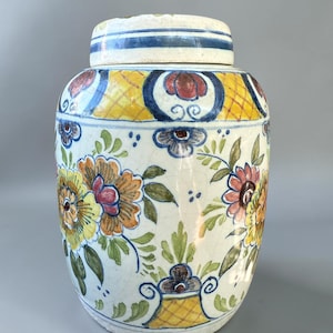 May include: A ceramic ginger jar with a lid, featuring a cream-colored base with a hand-painted floral pattern in yellow, orange, blue, and green. The design includes intricate lattice details, scrolling accents, and a visible crackle glaze finish.