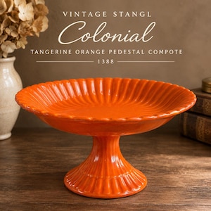 Könnte beinhalten: Eine mandarin-orangefarbene Fußschale mit geriffeltem, gewelltem Rand und passendem geriffeltem Fuß. Der Text VINTAGE STANGL Colonial TANGERINE ORANGE PEDESTAL COMPOTE 1388 ist über der Keramikschale mit glänzender Oberfläche zu sehen.