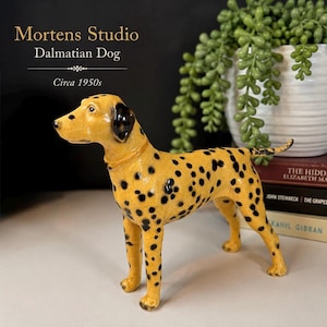 May include: A yellow ceramic Dalmatian dog figurine with black spots and a glossy finish. The text Mortens Studio Dalmatian Dog Circa 1950s is visible in the upper left corner. The dog stands in a profile position with its head turned slightly.