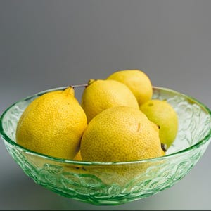 Puede incluir: Un cuenco de vidrio verde claro lleno de limones amarillos. El cuenco tiene un diseño decorativo. Los limones tienen piel texturizada y son un alimento saludable.