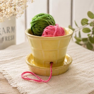 May include: A yellow ceramic yarn bowl with a textured design, holding green and pink yarn balls. The bowl sits on a matching saucer, with pink yarn extending from a hole. Ideal for knitting and crochet projects.