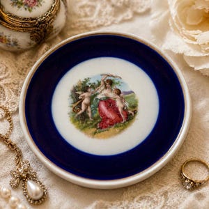 May include: A round porcelain saucer featuring a deep cobalt blue border and a central circular illustration of a figure in a red dress surrounded by two cherubs in a landscape setting, finished with a thin gold rim along the outer edge.