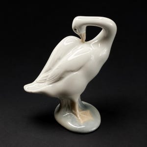 May include: A glossy white porcelain figurine of a goose in a preening pose. The bird has a curved neck, folded wings, and light tan coloring on its beak and webbed feet. It stands on a small, smooth, oval-shaped base with a soft gray tint.