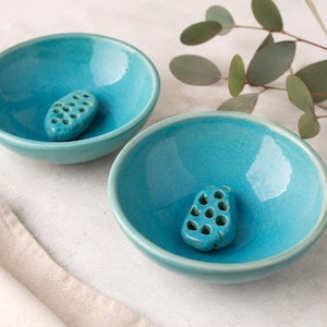 May include: Two turquoise ceramic bowls with a small, porous, turquoise stone inside each. The bowls are round with a smooth, glossy finish. A sprig of green leaves is in the background. A beige cloth is in the lower left corner.