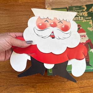May include: A vintage Eureka cardboard Santa Claus cutout featuring a white beard, red suit, and black boots, held together with metal staples. The cutout displays a classic illustration of Santa's face and torso, with a matching green packaging sleeve visible behind it.