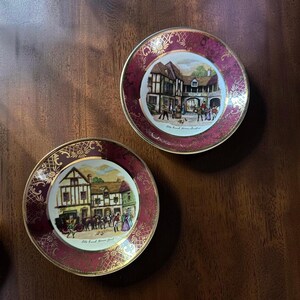 May include: Two round ceramic decorative plates with deep red borders featuring gold filigree patterns. The center of each plate displays a colorful illustration of a historic building with figures. Text on the plates reads Old Coach House Yard and Old Coach House Stratford.