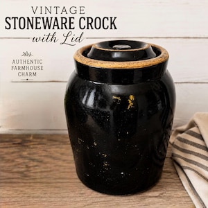 May include: A black stoneware crock with a matching lid, featuring a tan rim and distressed texture. The text VINTAGE STONEWARE CROCK with Lid and AUTHENTIC FARMHOUSE CHARM is visible above the object, which has a cylindrical body and a knobbed lid.