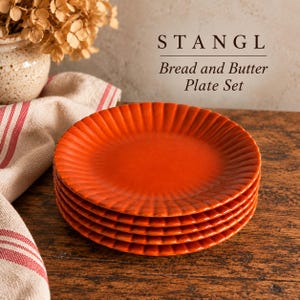Könnte beinhalten: Ein Stapel aus fünf runden, orangefarbenen Keramik-Brot- und Buttertellern mit geriffeltem, gewelltem Rand. Der Text STANGL Bread and Butter Plate Set ist über den Tellern zu sehen, die auf einer dunklen Holzoberfläche stehen.
