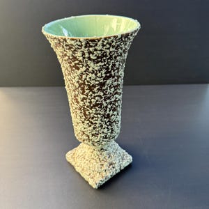 May include: A pedestal vase featuring a dark brown exterior with a textured, speckled light green finish. The interior of the vase is smooth and light green. The vessel has a wide, flared rim and sits on a square base with a matching texture.