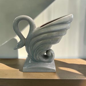 Puede incluir: Una maceta o jarrón de cerámica azul claro con forma de cisne y acabado brillante. El diseño presenta un asa en forma de cuello curvado, una abertura ancha en la parte superior y un cuerpo escalonado en forma de ala sobre una base cuadrada.