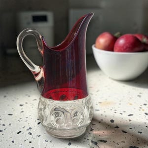 May include: A vintage glass pitcher with a ruby red upper section and a clear, patterned base. The pitcher has a curved handle and a spout. The pitcher is likely used for serving beverages. The pitcher is sitting on a speckled countertop.
