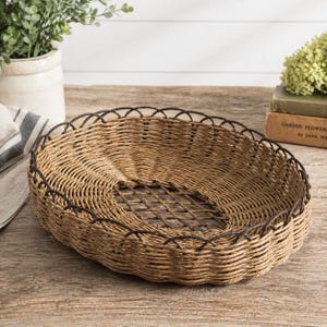 May include: Oval woven basket with a dark brown metal trim. The basket is made of light brown woven material and has a decorative scalloped edge. The center of the basket has a grid pattern.